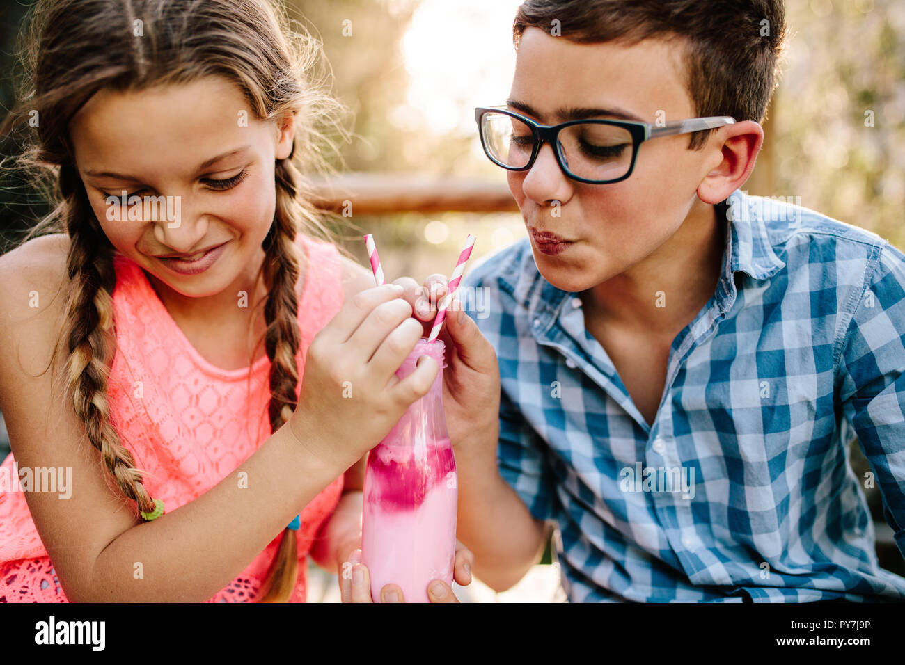 Close up of kids in love sitting outdoors drinking a smoothie with two straws. Smiling boy and girl sharing a milk shake sitting outdoors. Stock Photo