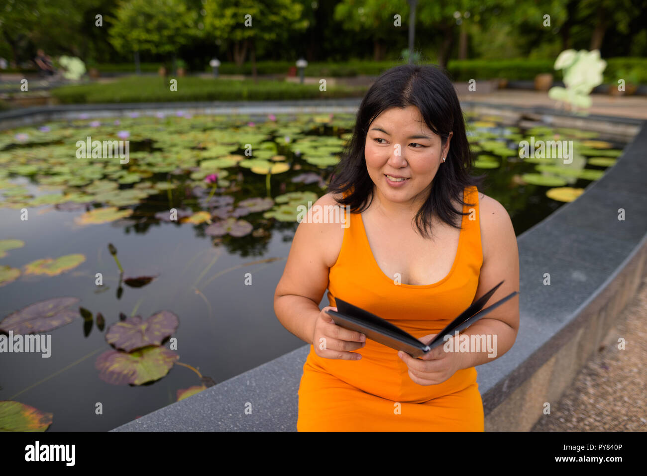 Beautiful overweight Asian woman smiling and reading book in park Stock Photo