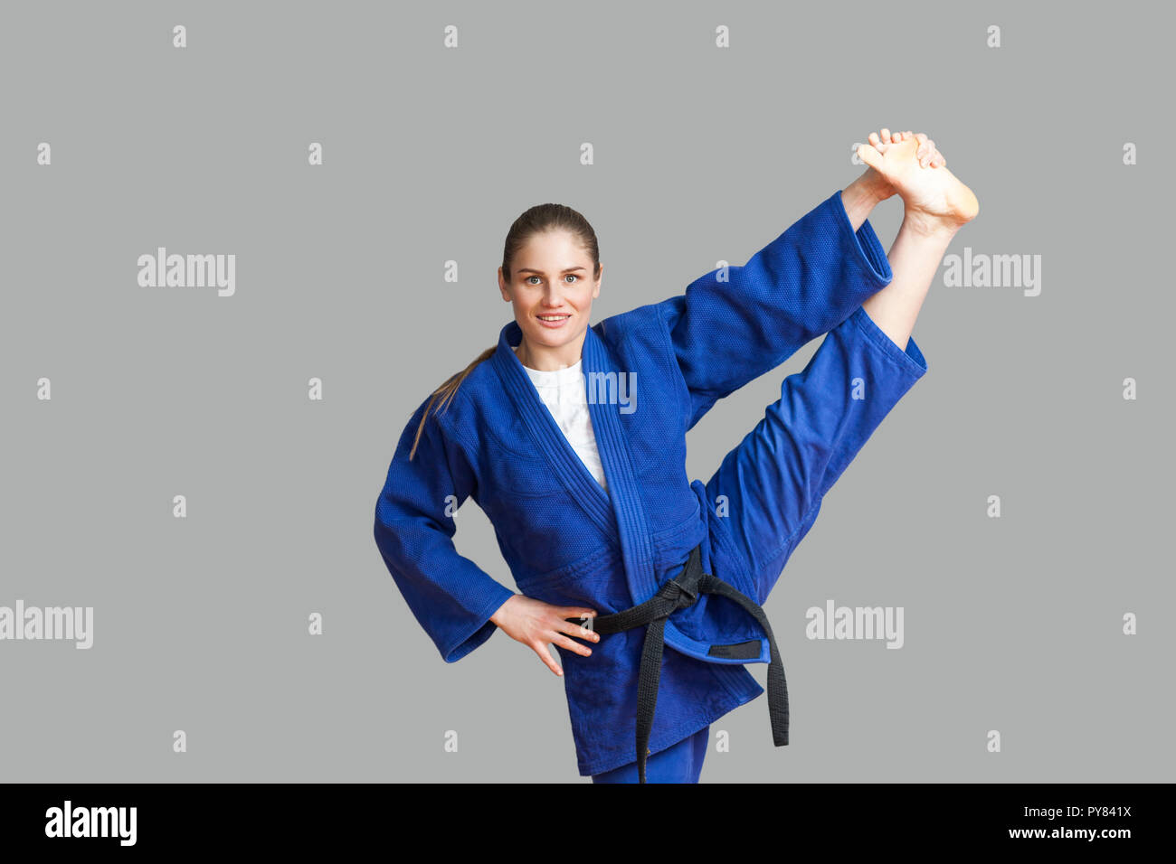 Beautiful happy athletic karate woman in blue kimono with black belt making vertical twine and looking at camera with toothy smile. Japanese martial a Stock Photo