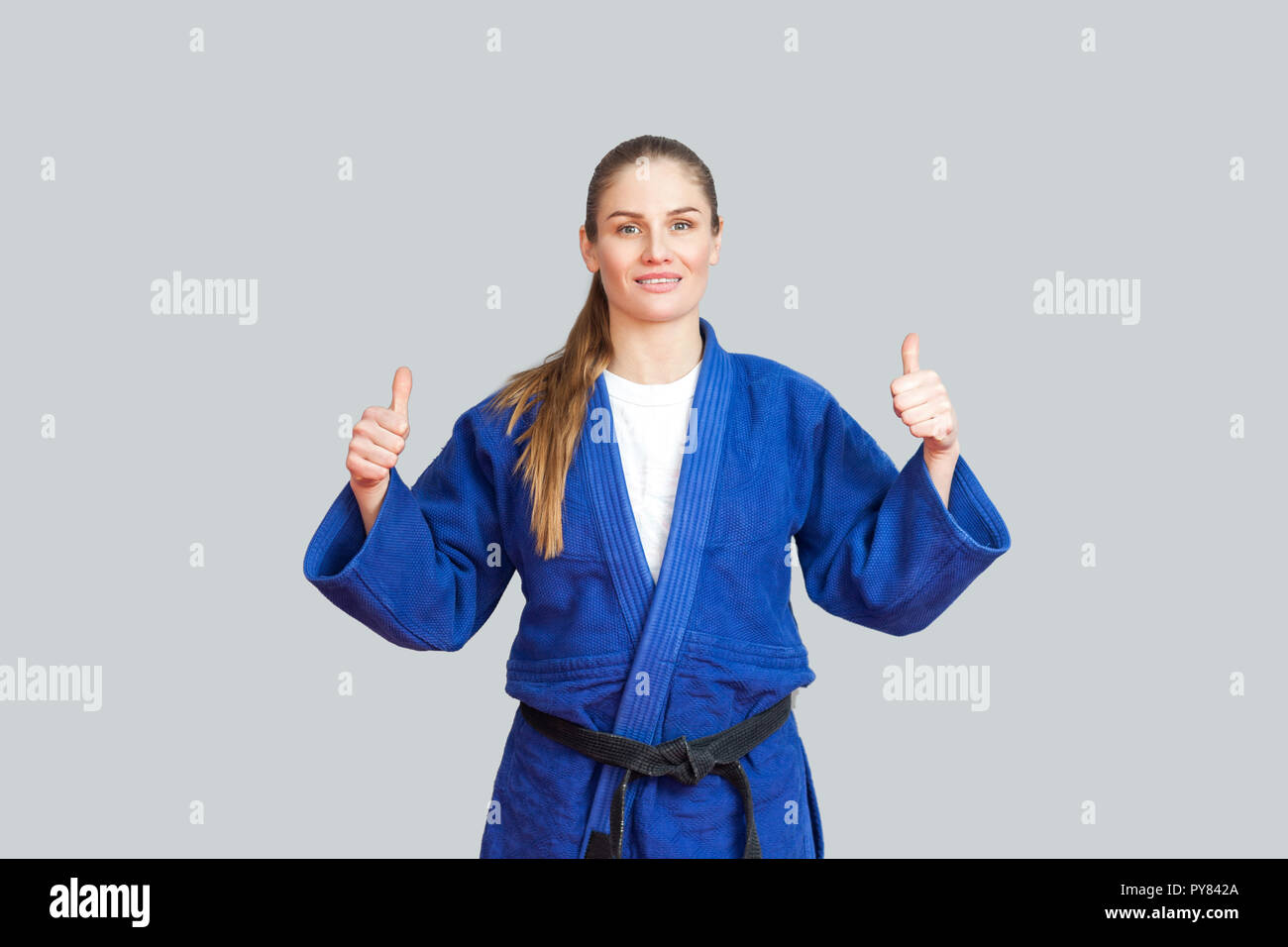 Positive athletic karate woman in blue kimono with black belt standing, showing thumbs up and looking at camera with toothy smile. Japanese martial ar Stock Photo