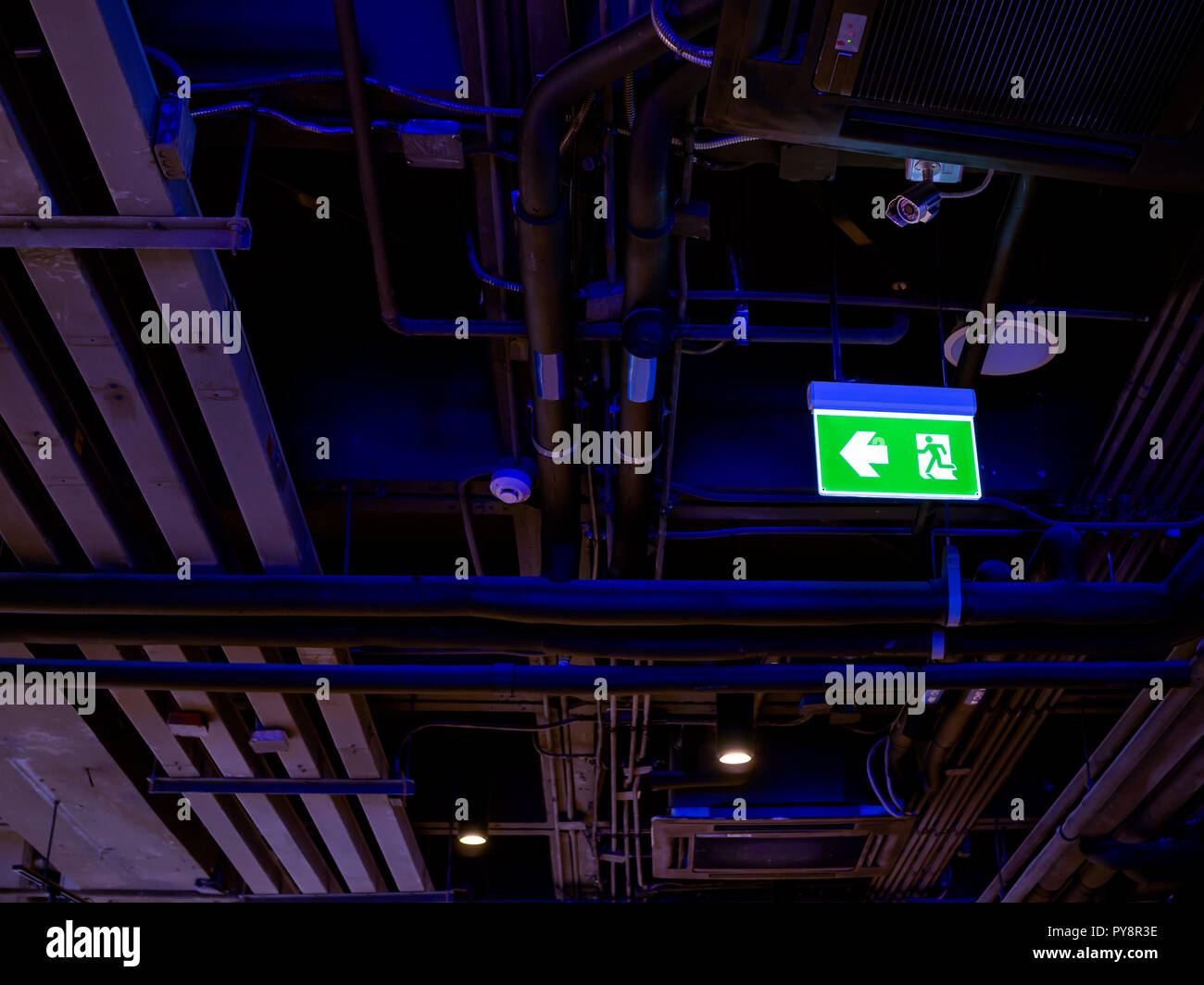 Illuminated green fire exit LED light sign on the ceiling in the dark with pipe line system inside the building. Stock Photo