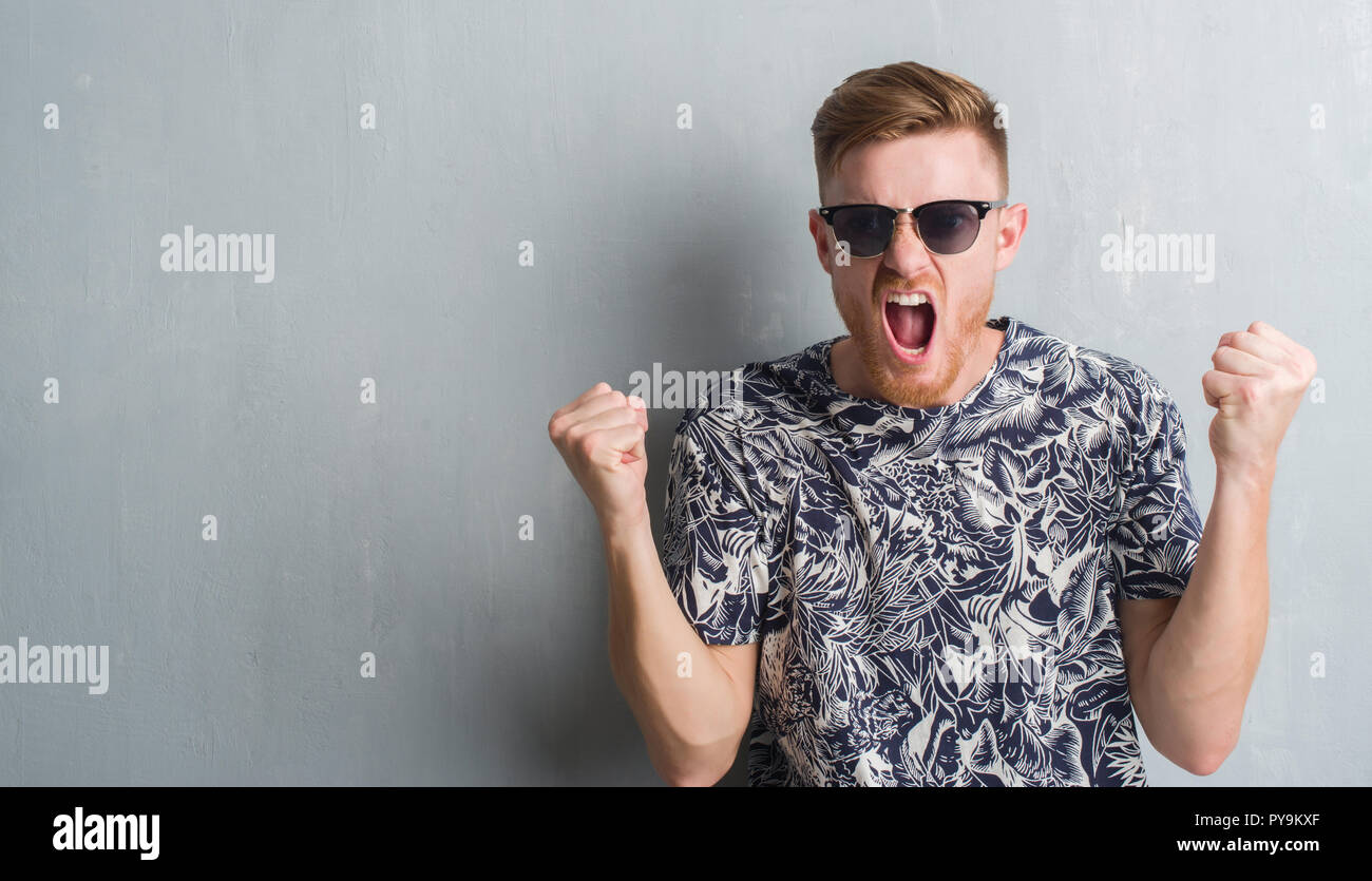 Young redhead man over grey grunge wall wearing sunglasses annoyed and ...