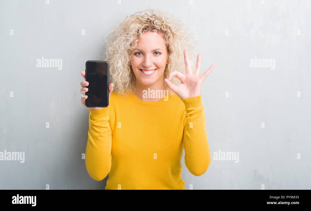 Young blonde woman over grunge grey background showing blank screen of smartphone doing ok sign with fingers, excellent symbol Stock Photo