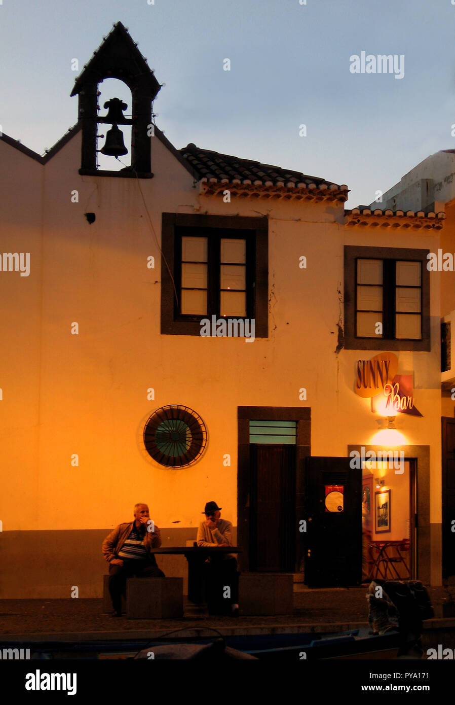 Two mature men sit outside The Sunny Bar in Funchal, Madeira, in the early evening as the light fades and the street lights come on. Above them is a small bell tower Stock Photo