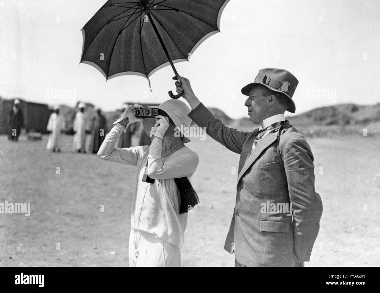 Taking pictures in the 1930s. A woman is photographing something with a Stereo camera. A man is standing by her side holding an umbrella to block the sun. 1930s Stock Photo