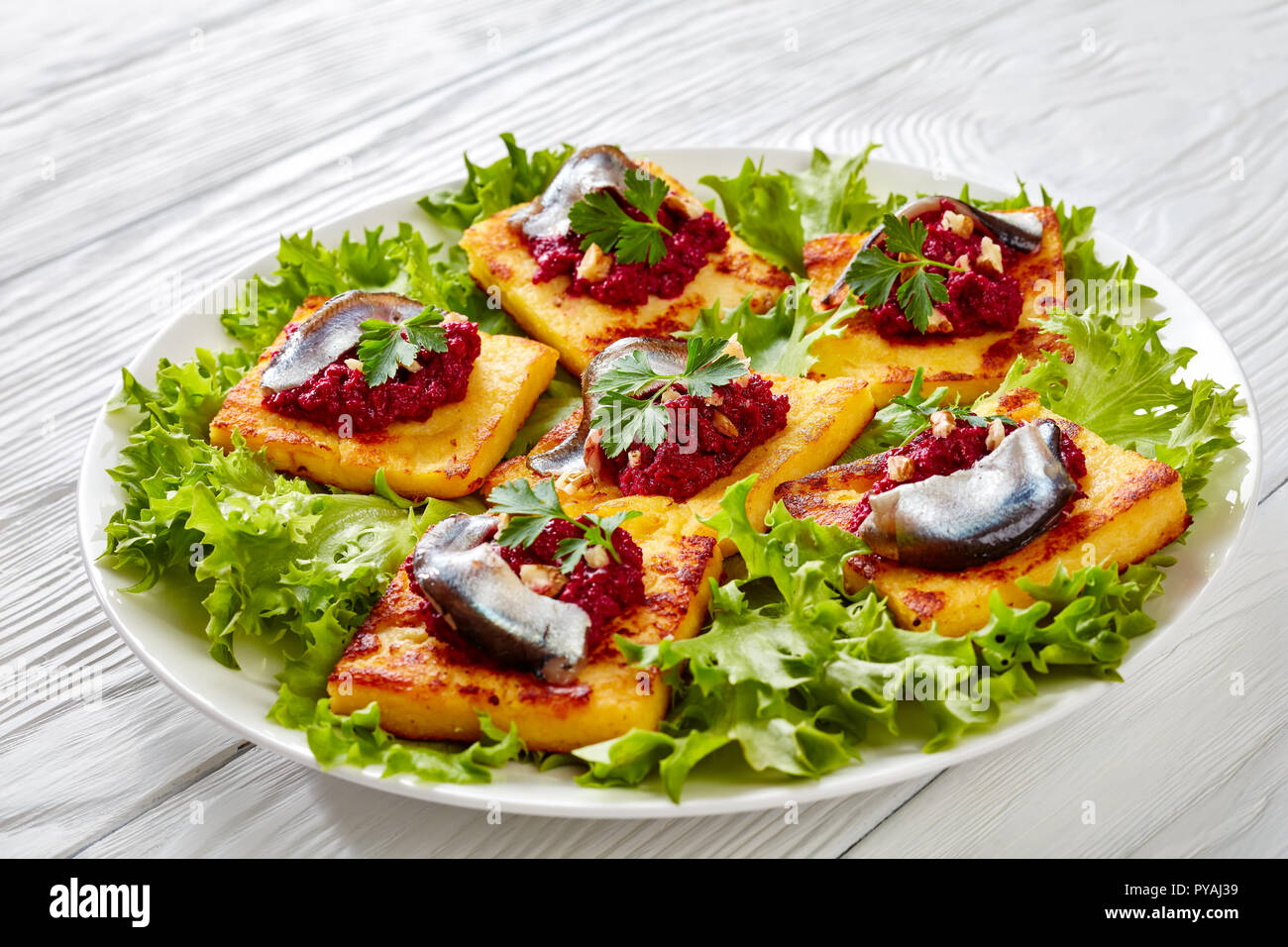 crispy Fried Polenta Squares with Creamy beetroot puree, topped with anchovies and garnished with fresh parsley on a white plate on a wooden table, ho Stock Photo
