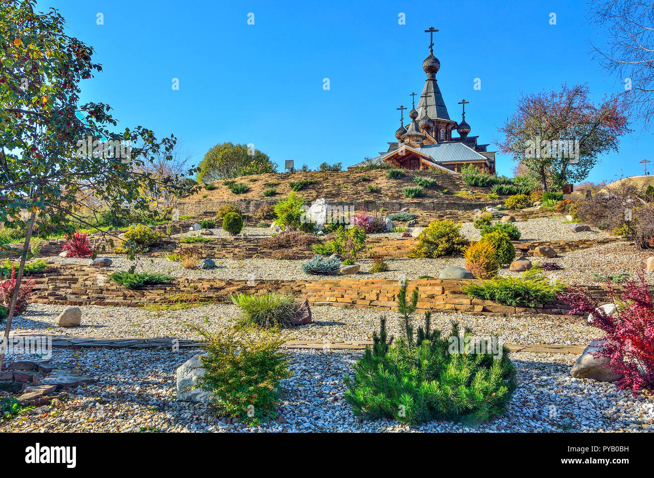 Dwarf trees, bushes and flowers on the stony terraces fortified by gabions on hill slope - front yard of orthodox Temple. Domes and crosses of church  Stock Photo
