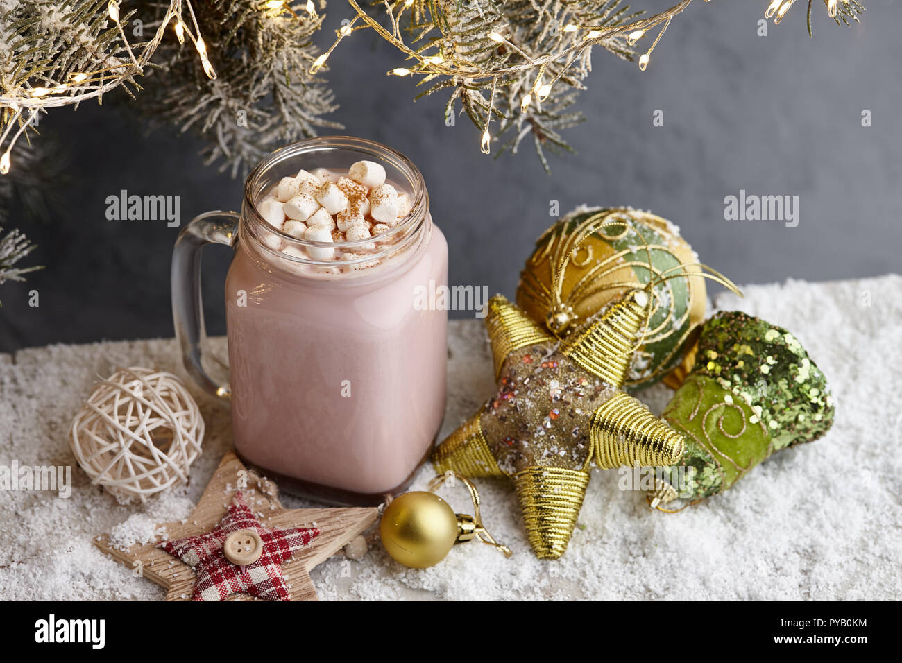 Glass winter cup of hot chocolate drink with marshmallow and Christmas toys on snowy surface under a fir tree with lights Stock Photo