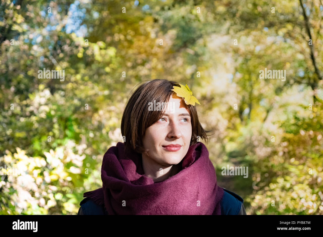 Portrait of young woman in scarf with autumn leave on her head. Maple leaf on the hair of a female person standing in beautiful green and yellow natur Stock Photo