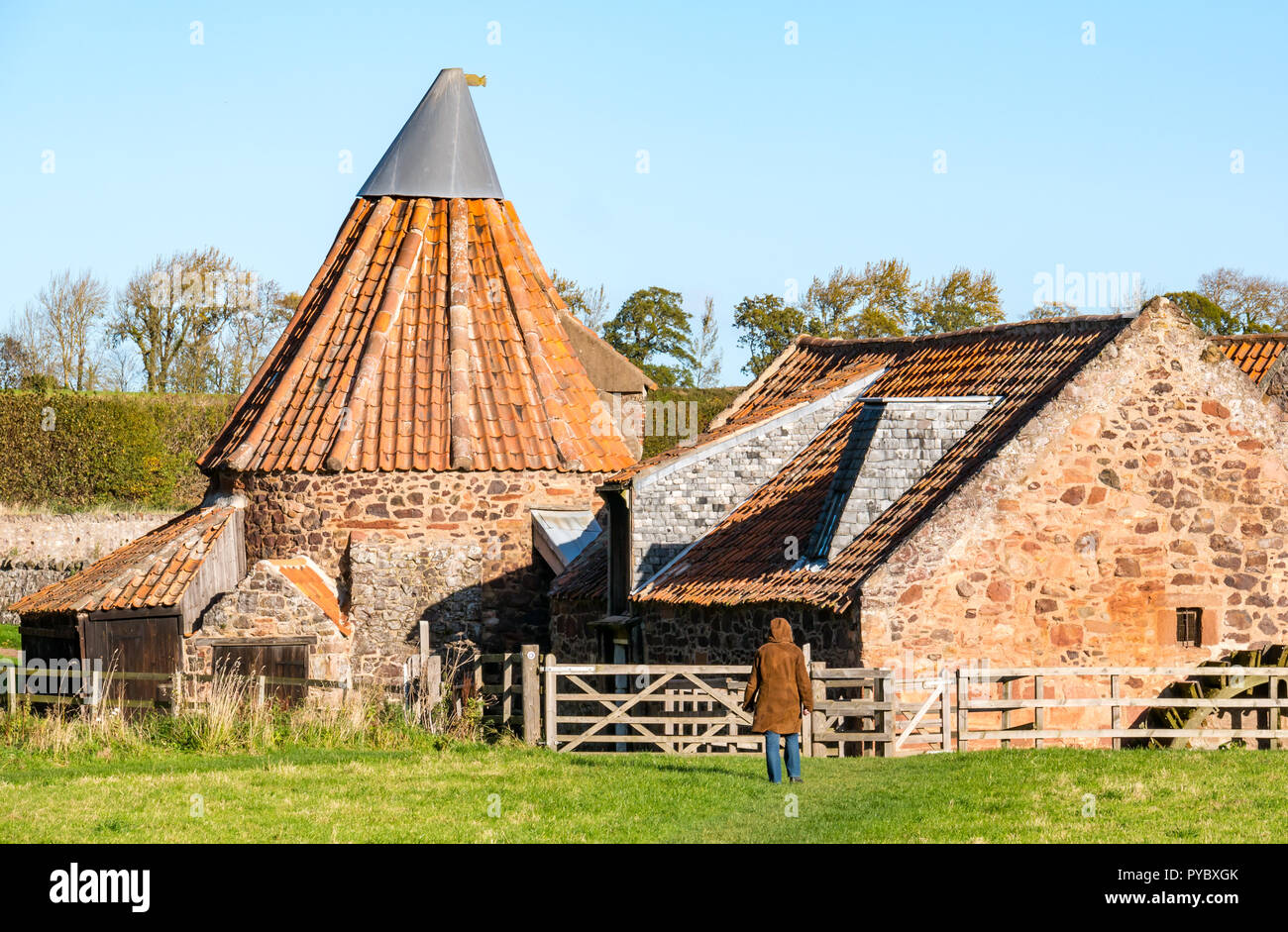 East Linton, East Lothian, Scotland, United Kingdom, 27th October 2018. UK Weather: A beautiful cold Autumnal sunny day lights up the quirky 18th century watermill, Preston Mill. A woman walks towards the old mill gate across a grass field Stock Photo