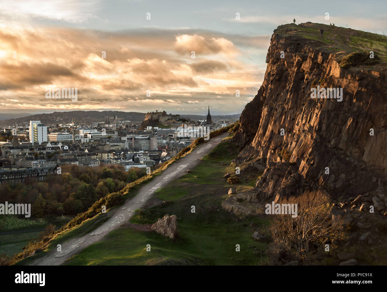 View of the Salisbury Crags at Holyrood Park, in Edinburgh, with the Castle in the background, and evening light on the face of the cliffs and clouds Stock Photo