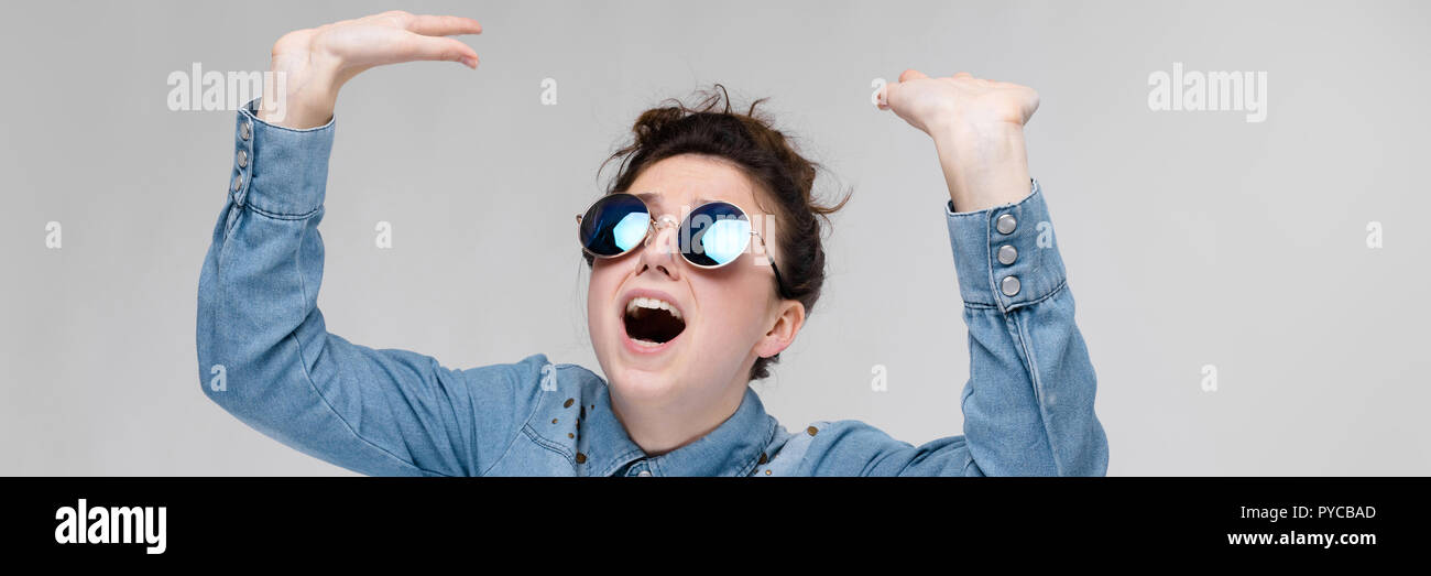 Young brunette girl in round glasses. Hairs are gathered in a bun. The girl lifted her hand up. Stock Photo