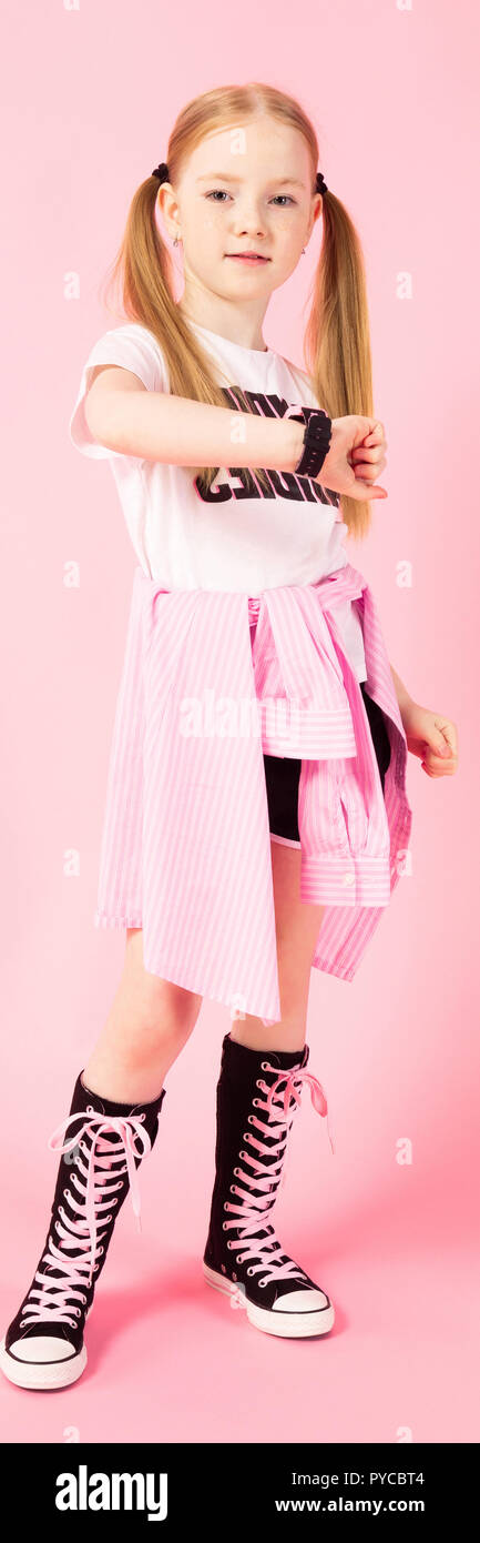 Girl with red tails on a pink background. A charming girl in bright sports clothes looks at her watch. Stock Photo