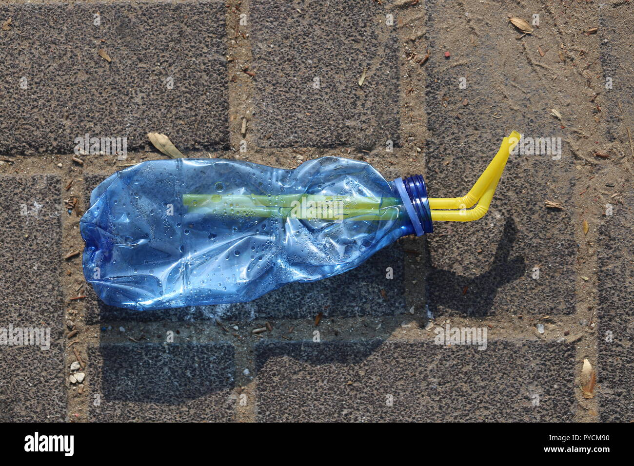 Crushed Plastic Bottle on the Sidewalk. Blue squeezed plastic bottle with two drinking straws inside, on the pavement. Disposable bottle thrown away. Stock Photo