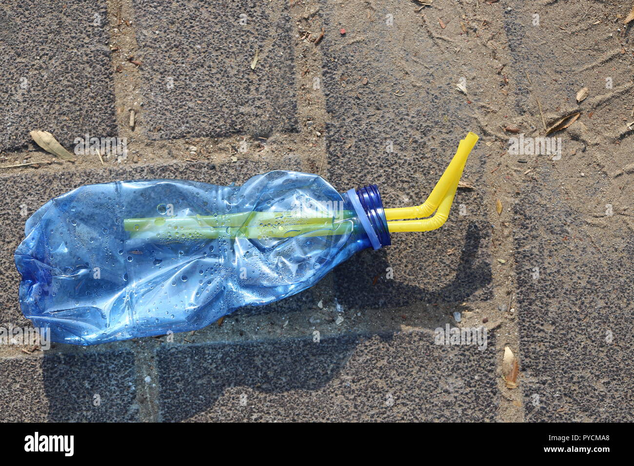 Crushed Plastic Bottle on the Sidewalk. Blue squeezed plastic bottle with two drinking straws inside, on the pavement. Disposable bottle thrown away Stock Photo