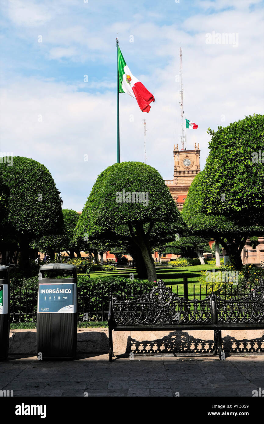 Two Mexican flags foregrounded by artificially manicured trees and a  trash bin for "inorganic" waste; symbolic of the inorganic nature of the nation. Stock Photo