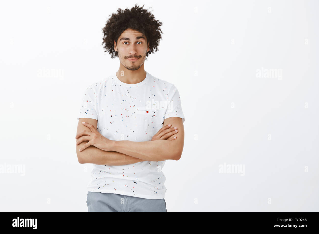 Man having bet with friend, being confident he win. Portrait of attractive carefree hispanic guy with tanned skin and curly hair, holding hands crossed with self-assured smile standing over grey wall Stock Photo