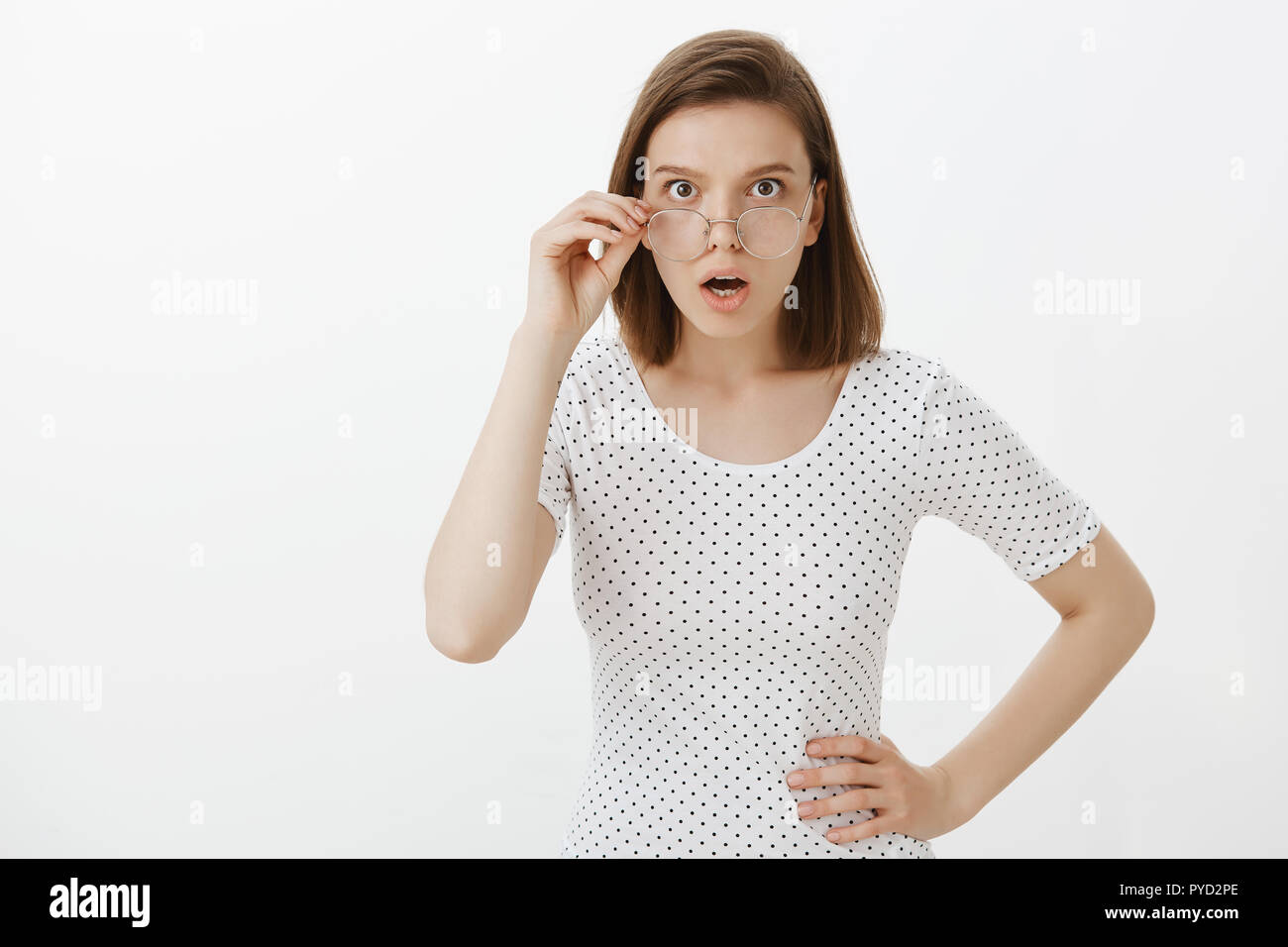 Shocked intense caucasian girl, holding hand on hip, taking off glasses, dropping jaw and gasping, being questioned and confused with strange situation, standing puzzled over gray background Stock Photo