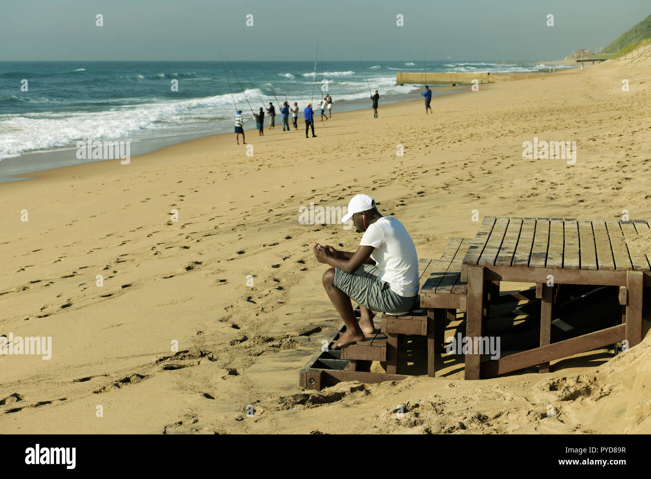 Durban, KwaZulu-Natal, single young adult male looking at mobile phone, sitting on steps of beach stairs where fishermen are fishing, The Bluff Stock Photo