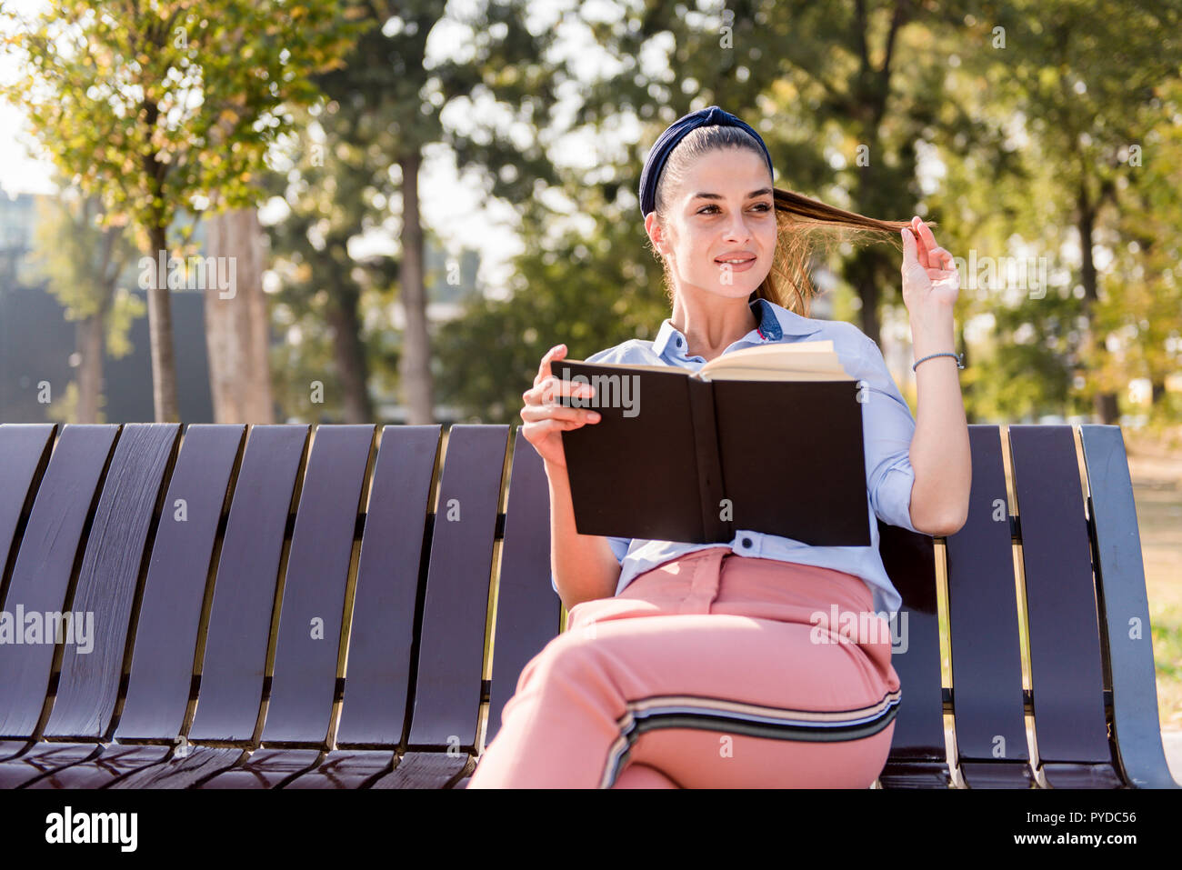 Cheerfull young woman holding open book and reading on bench in autumn ...