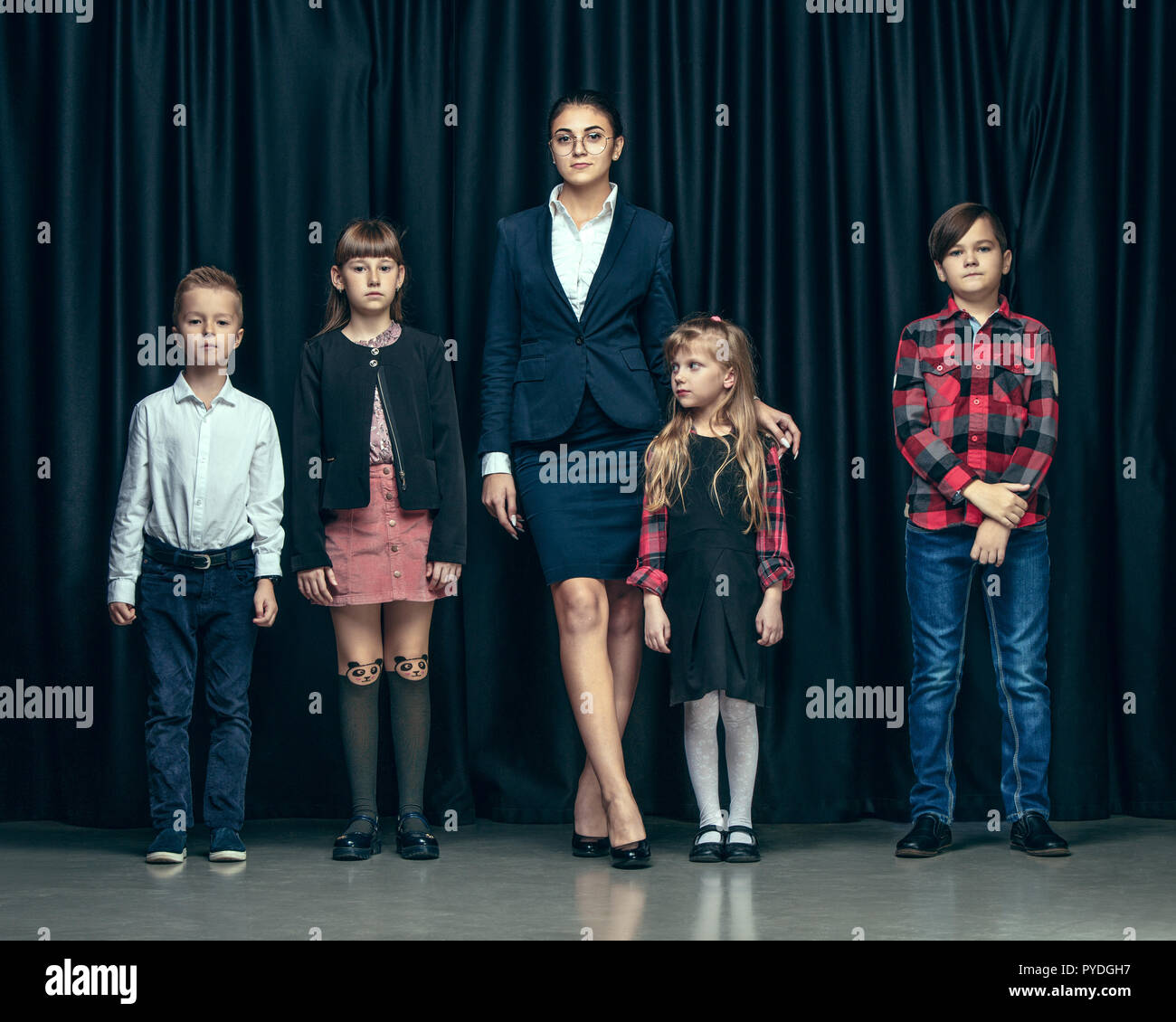 Cute smiling happy stylish children and female teacher on dark background.  Beautiful stylish teen girls and boy standing together and posing on the school  stage in front of the curtain. Classic style.