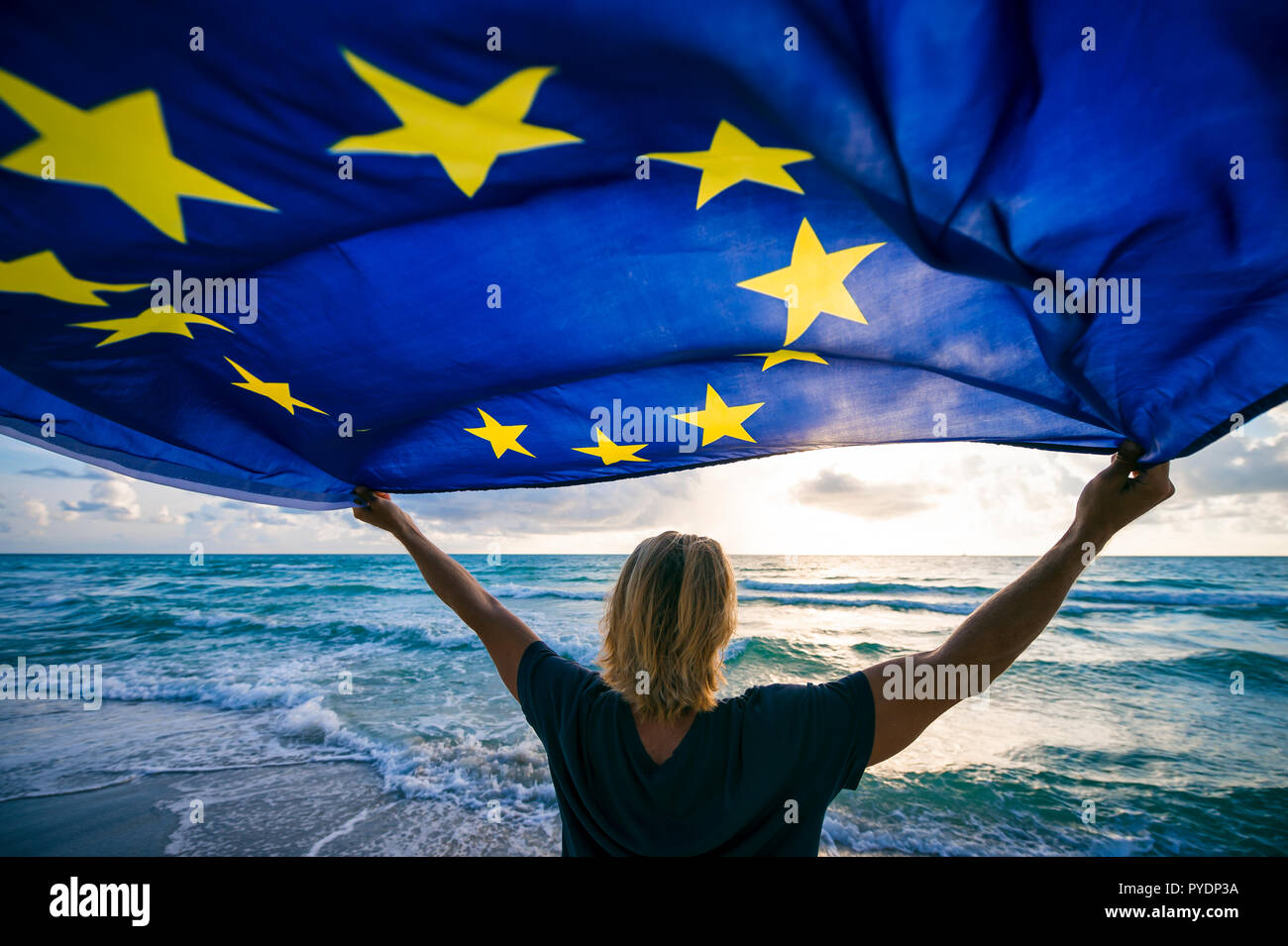 Man holding EU European Union flag waving in front of Mediterranean sunrise seascape Stock Photo