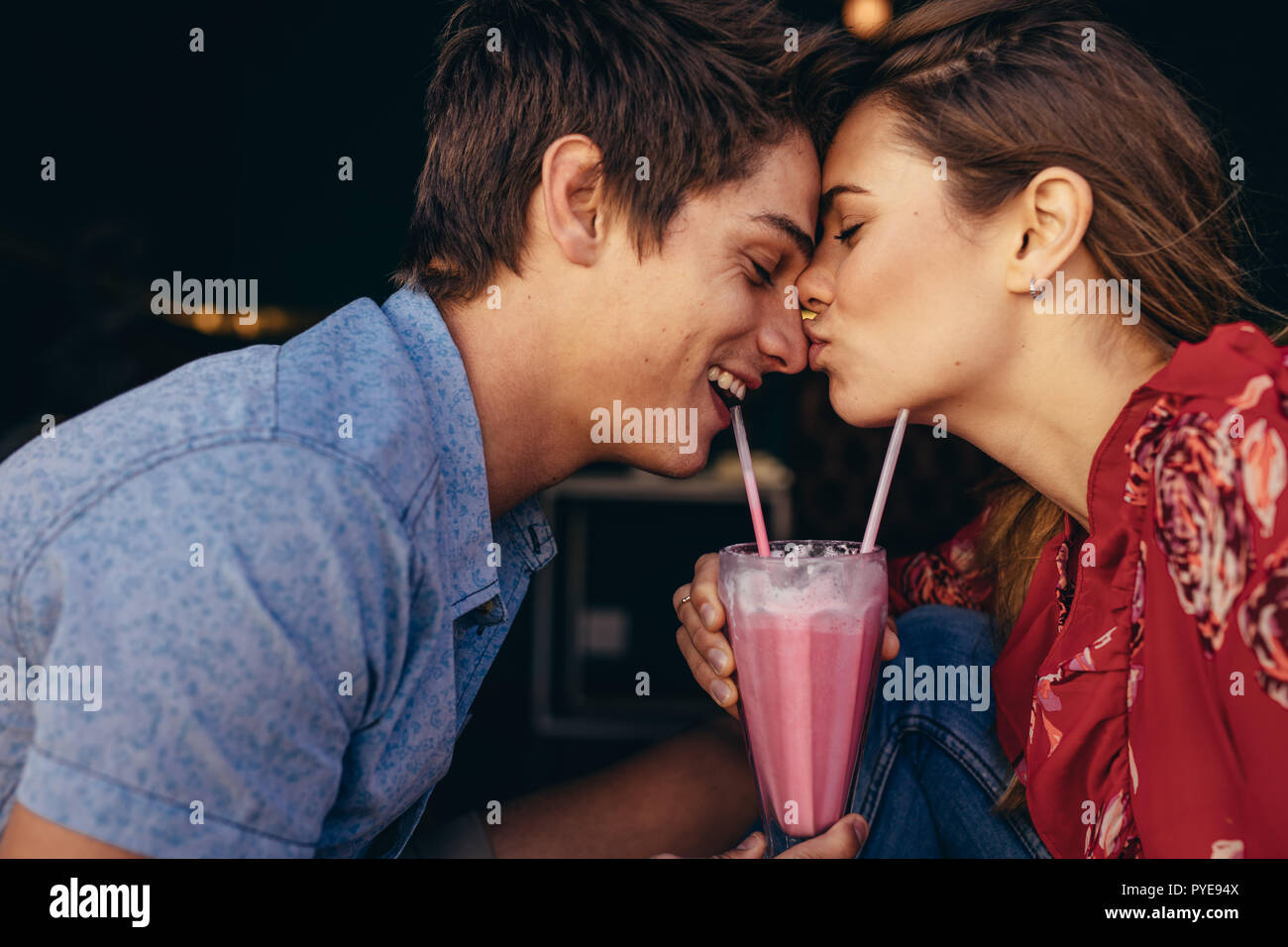 Woman kissing her boyfriend on his nose while on date at a restaurant. Couple at a restaurant in romantic mood sharing a milkshake. Stock Photo