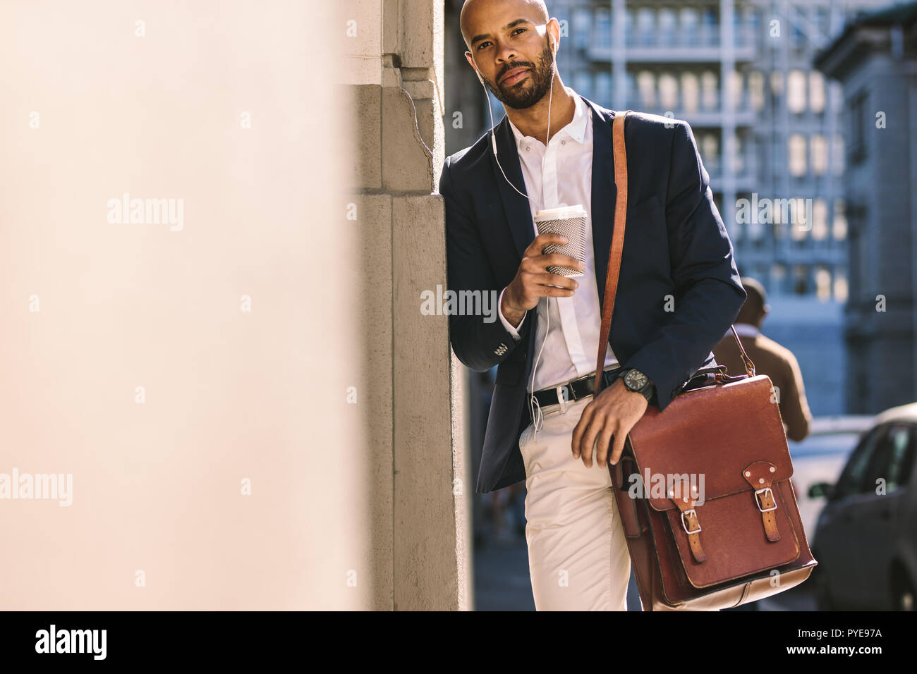 Portrait of handsome young man leaning to wall outdoors with coffee. African businessman with handbag wearing earphones and holding a cup of coffee. Stock Photo