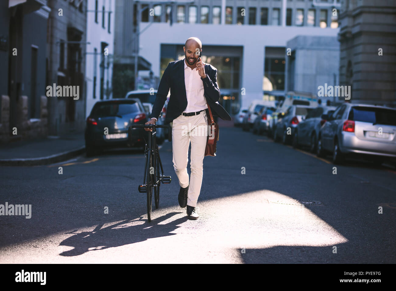 Handsome young african man wearing suit talking on phone while walking with bicycle to work in the morning. Businessman going to work with his bicycle Stock Photo