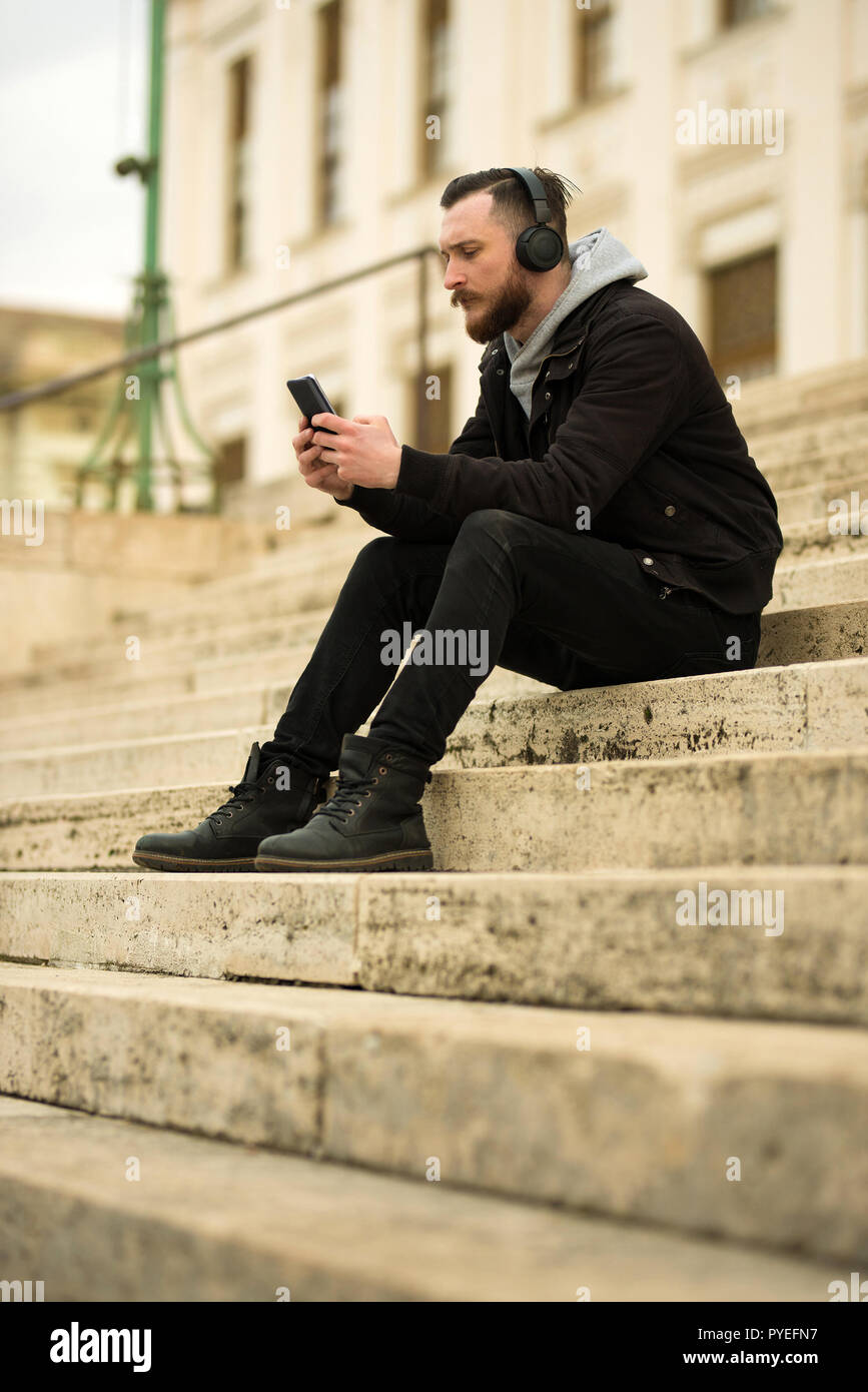 A handsome young hipster man sitting on the stairs and checking his ...