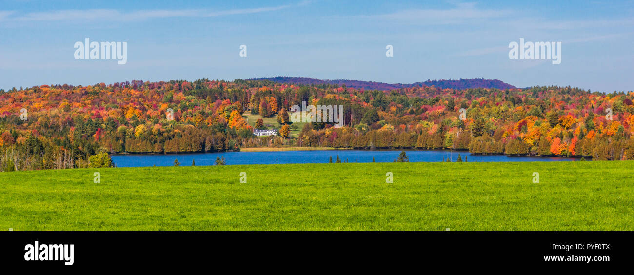 panorama banner of white house in the country  with blue pond, green fields and  trees  dressed in bright autumn colors of fall foliage Stock Photo