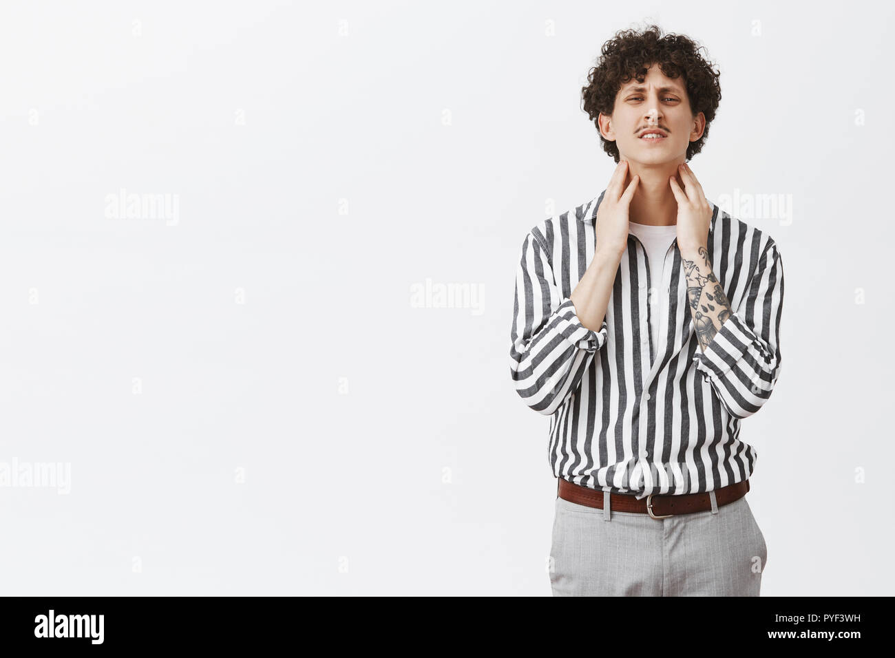 Man feeling pain in throat might getting cold. Uncomfortable curly-haired guy with moustache and tattoos in striped shirt raising head clenching teeth and frowning touching neck having discomfort Stock Photo