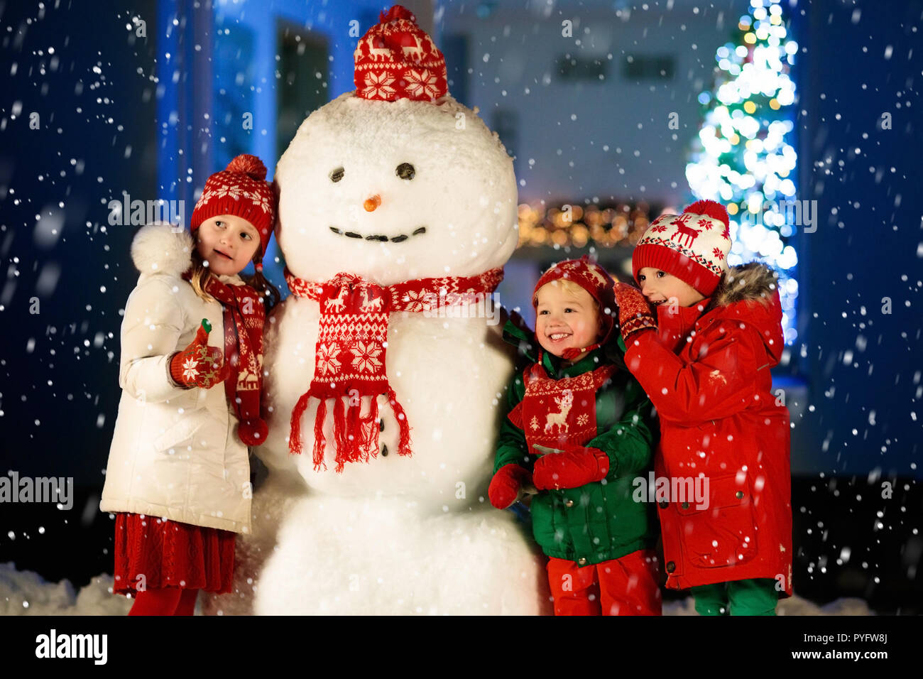 Kids build snowman on Christmas eve. Children in snowy backyard next to window to living room with Christmas tree and decorated fireplace. Boy and gir Stock Photo