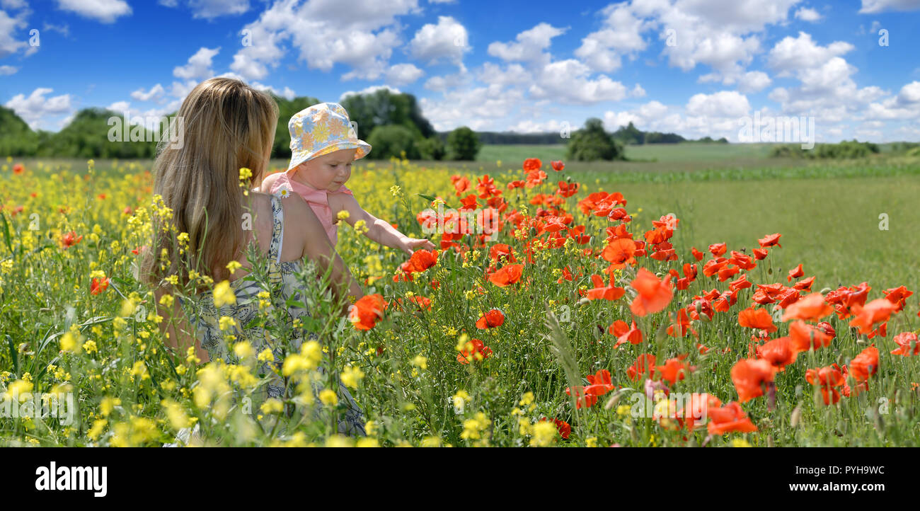 Young woman with a little child sitting on the flowery meadow, blue sky and white clouds in the background, panorama Stock Photo