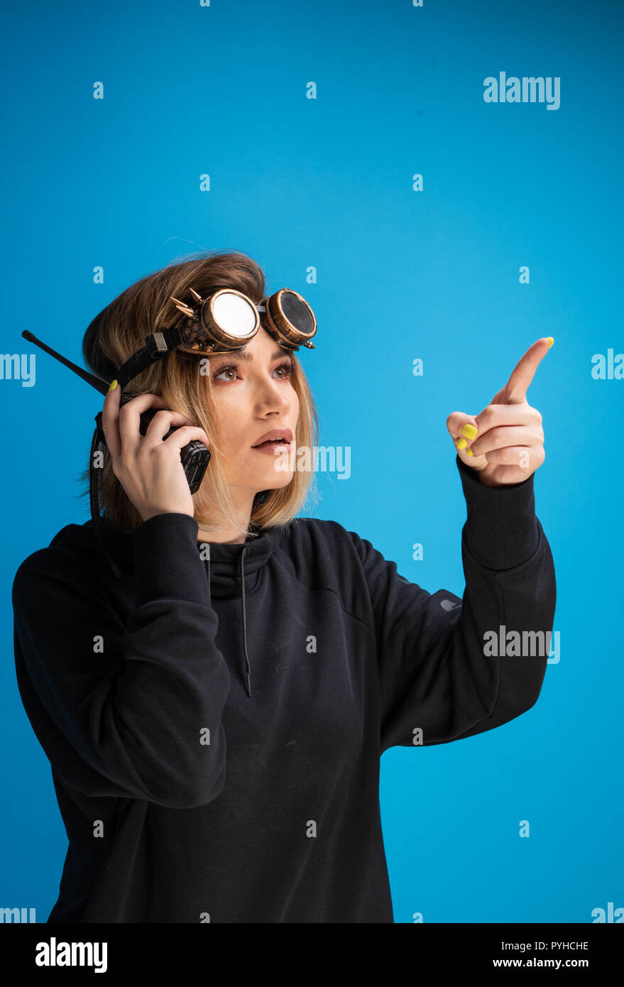 Portrait of dark blonde girl with steam punk glasses using a walkie talkie communication device and pointing with finger to the right side up Stock Photo