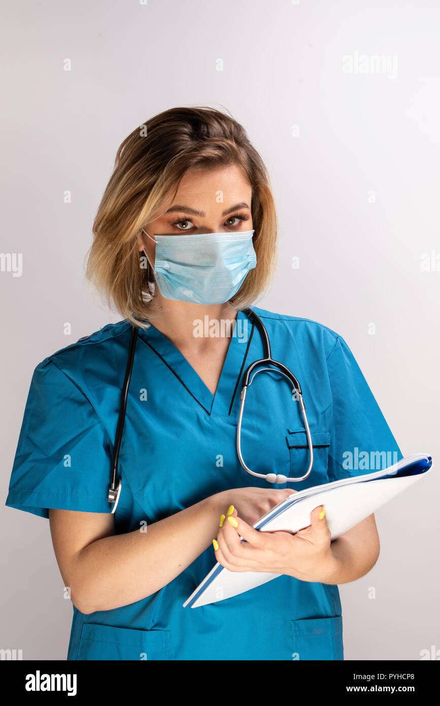 Portrait of young doctor with stethoscope wearing face mask holding paper results. Blue costume isolated against white background Stock Photo