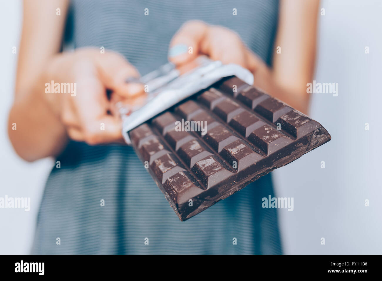 Female's hands holding unwrapped dark chocolate bar, close-up. Unrecognizable young woman standing and offering cocoa dessert. Stock Photo