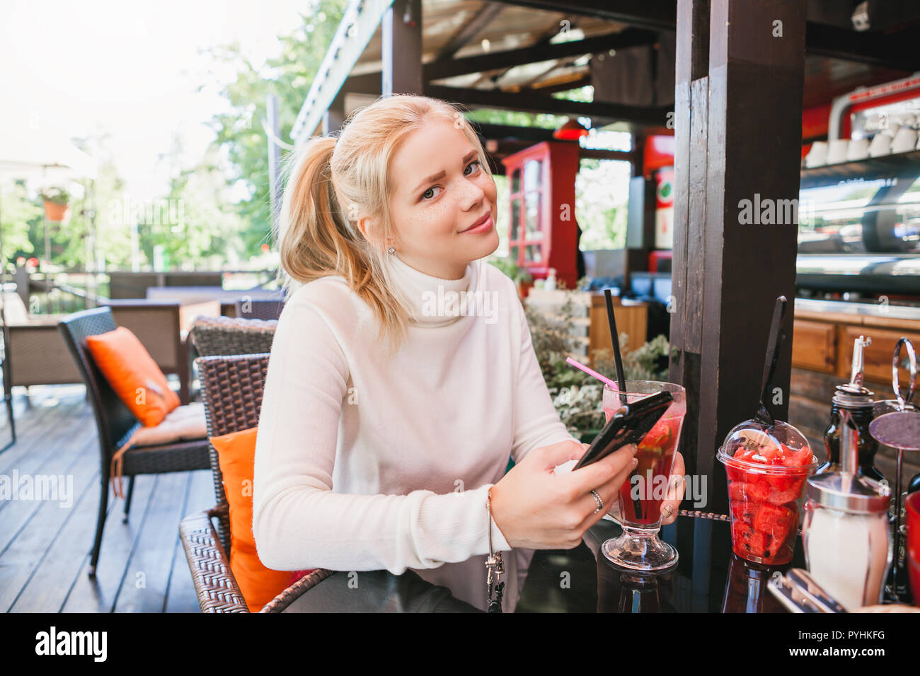 young girl with freckles texting message on her smartphone while drinking coctail with berries and mint in the outdoor restaurant Stock Photo