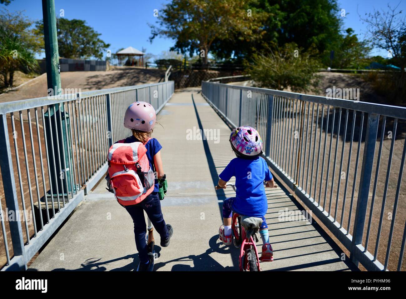 Young children riding a bike and a scooter across a pedestrian bridge