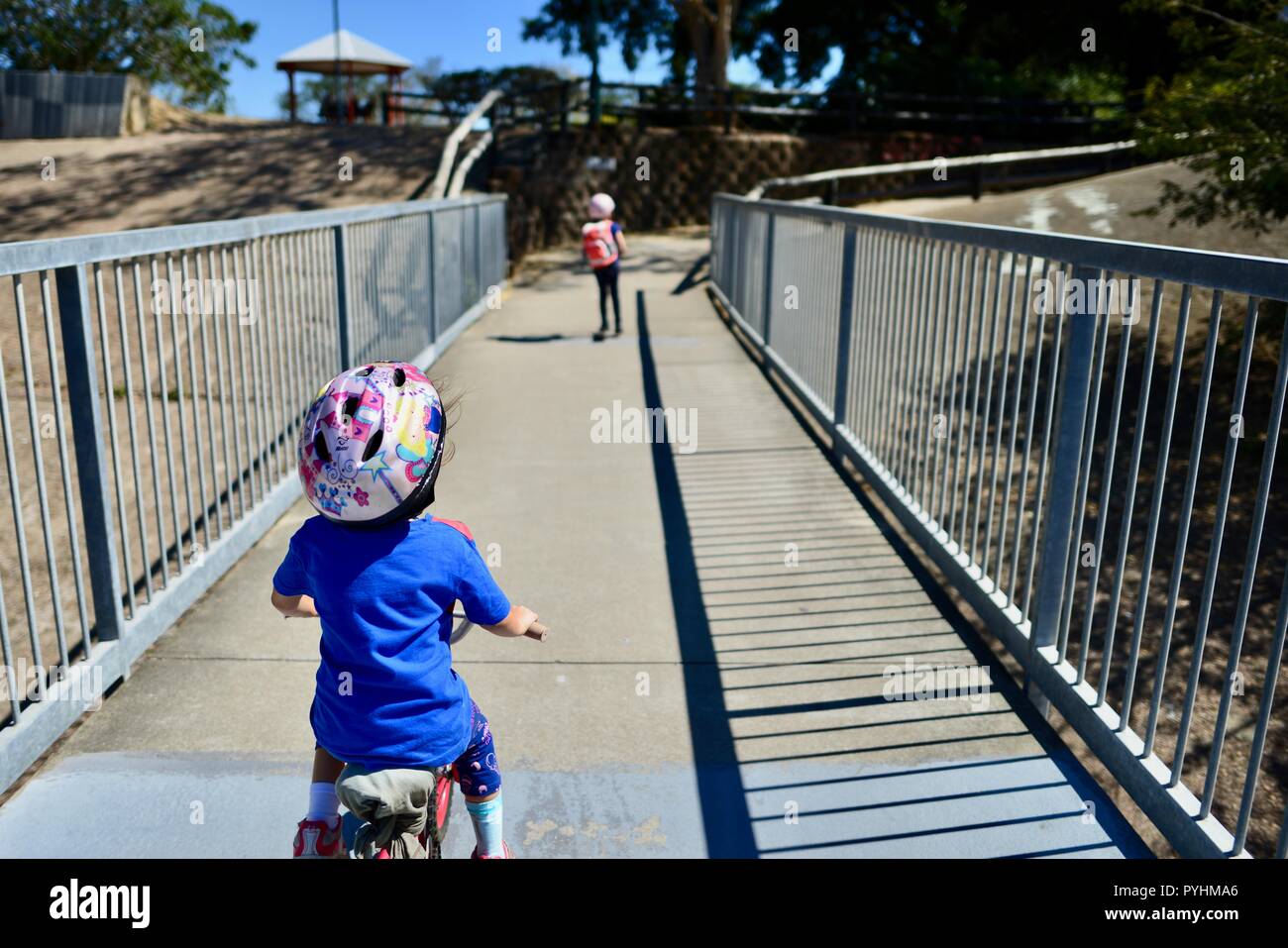Young children riding a bike and a scooter across a pedestrian bridge