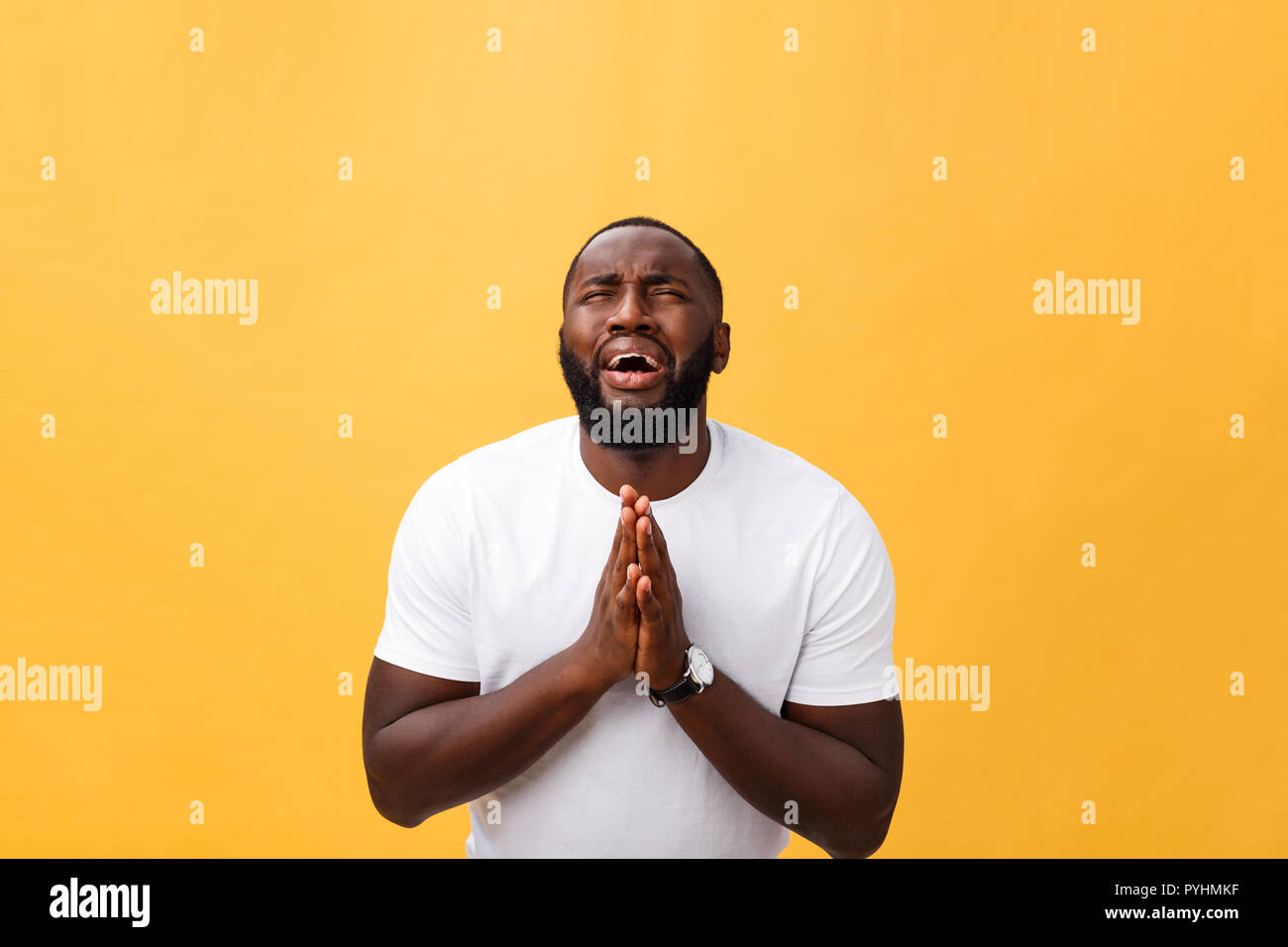 Studio portrait of young African American man in white shirt, holding hands in prayer, looking at the camera with thoughtful skeptical expression on his face, suspecting of something. Body language. Stock Photo
