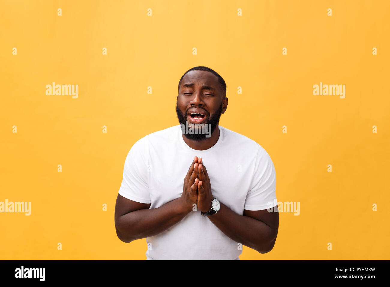 Studio portrait of young African American man in white shirt, holding hands in prayer, looking at the camera with thoughtful skeptical expression on his face, suspecting of something. Body language. Stock Photo