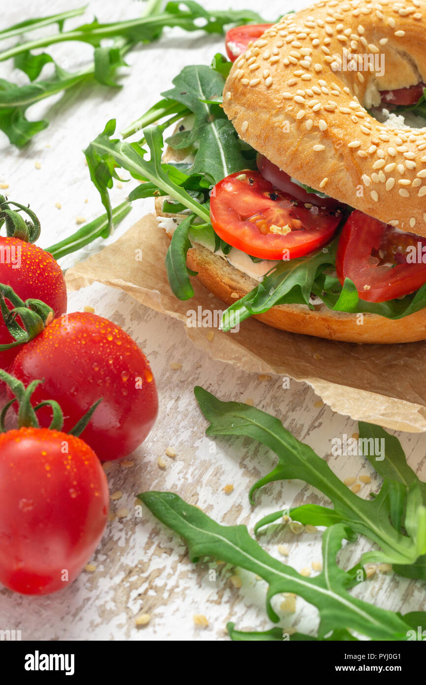 Frischer Sesambagel auf Backpapier mit Frischkäse, Tomaten und Rucola auf weißem Holzuntergrund Stock Photo