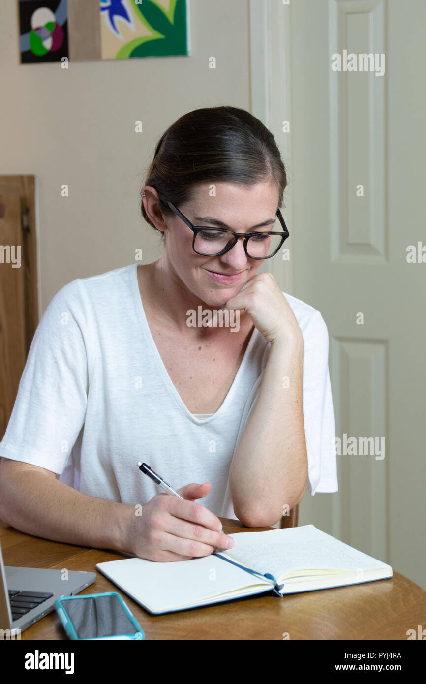Young woman artist/painter reading journal with cell phone and laptop resting chin on hand close up sitting at dining room table. Stock Photo