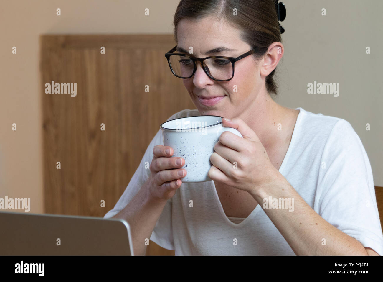 Young woman wearing glasses looking down at laptop holding a mug to take a drink. Close up of her face and hands and corner of the computer. Stock Photo