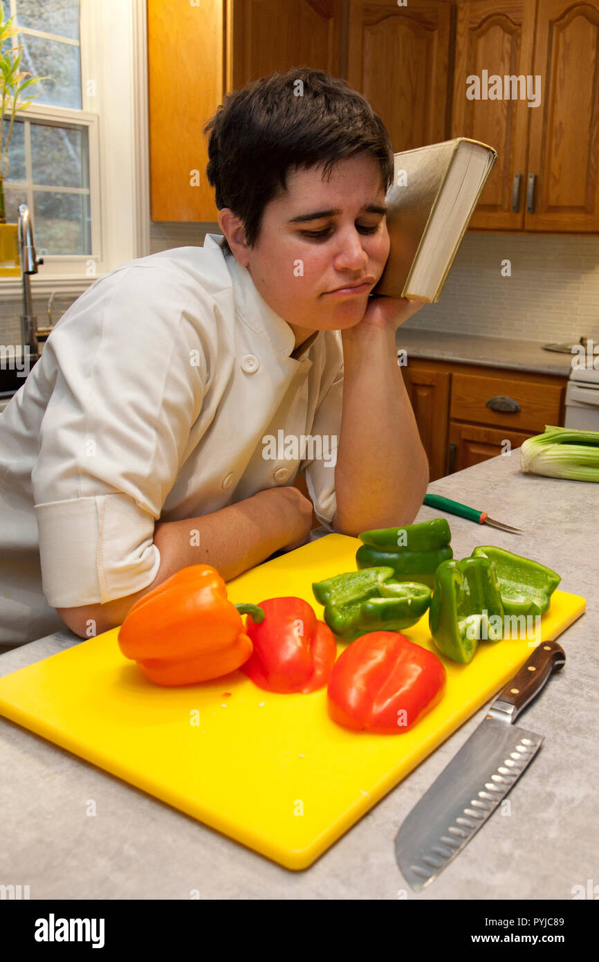 Dressed in a chef's outfit a person leans their head against a cookbook in despair with vegetables in the foreground Stock Photo