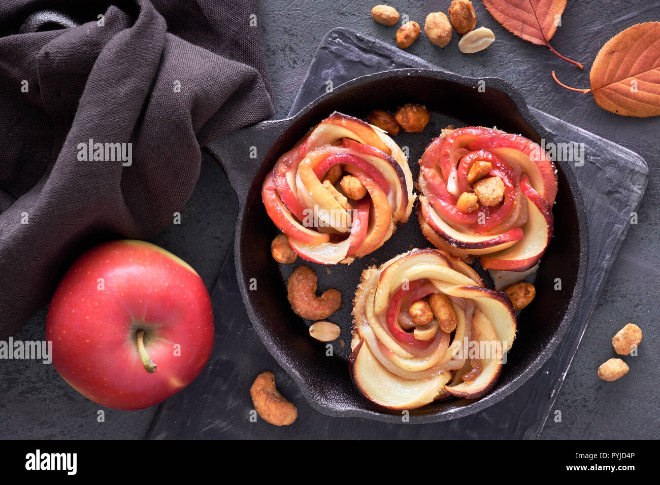 Three puff pastries with rose shaped apple slices baked in cast iron skillet and a fersh red apple. Top lay on dark background with linen towel and ca Stock Photo