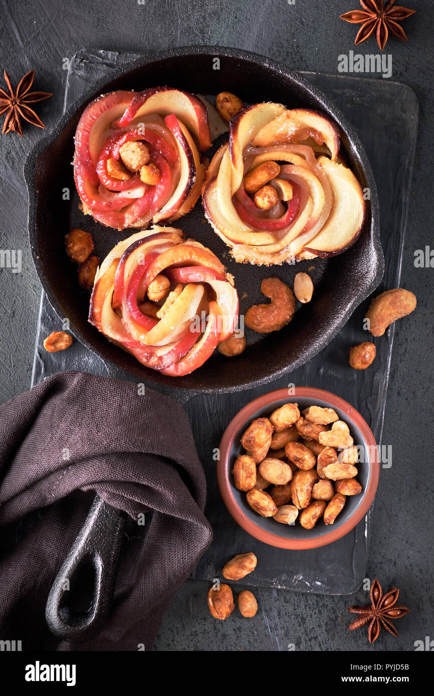 Three homemade puff pastries with rose shaped apple slices baked in cast iron skillet. Top lay on dark background with linen towel and caramelized nut Stock Photo