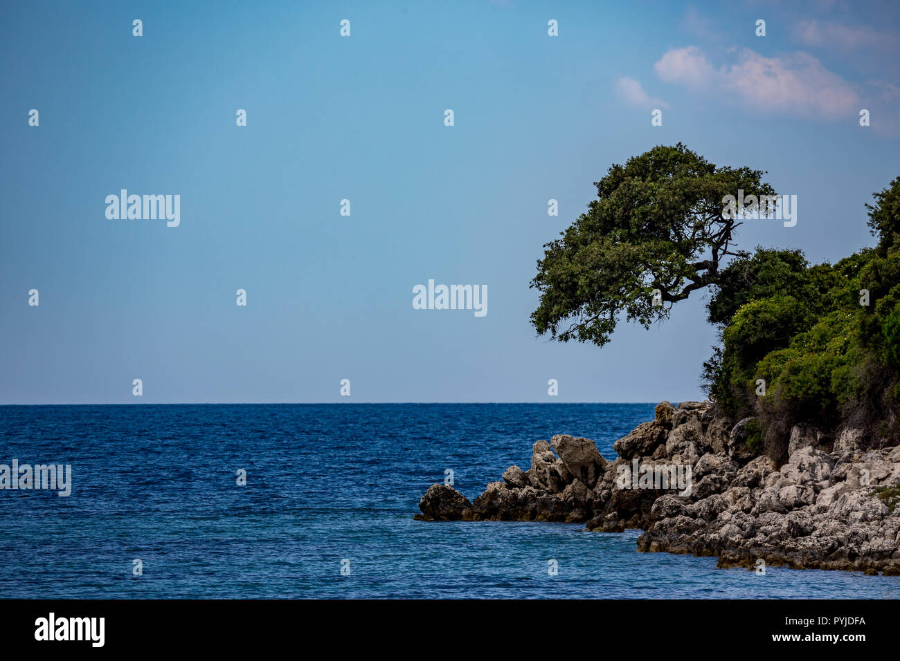 Spring daytime beautiful Ionian Sea with clear turquoise water, rocks and trees coast view from Ksamil beach, Albania. Deep blue sky with white clouds. Stock Photo