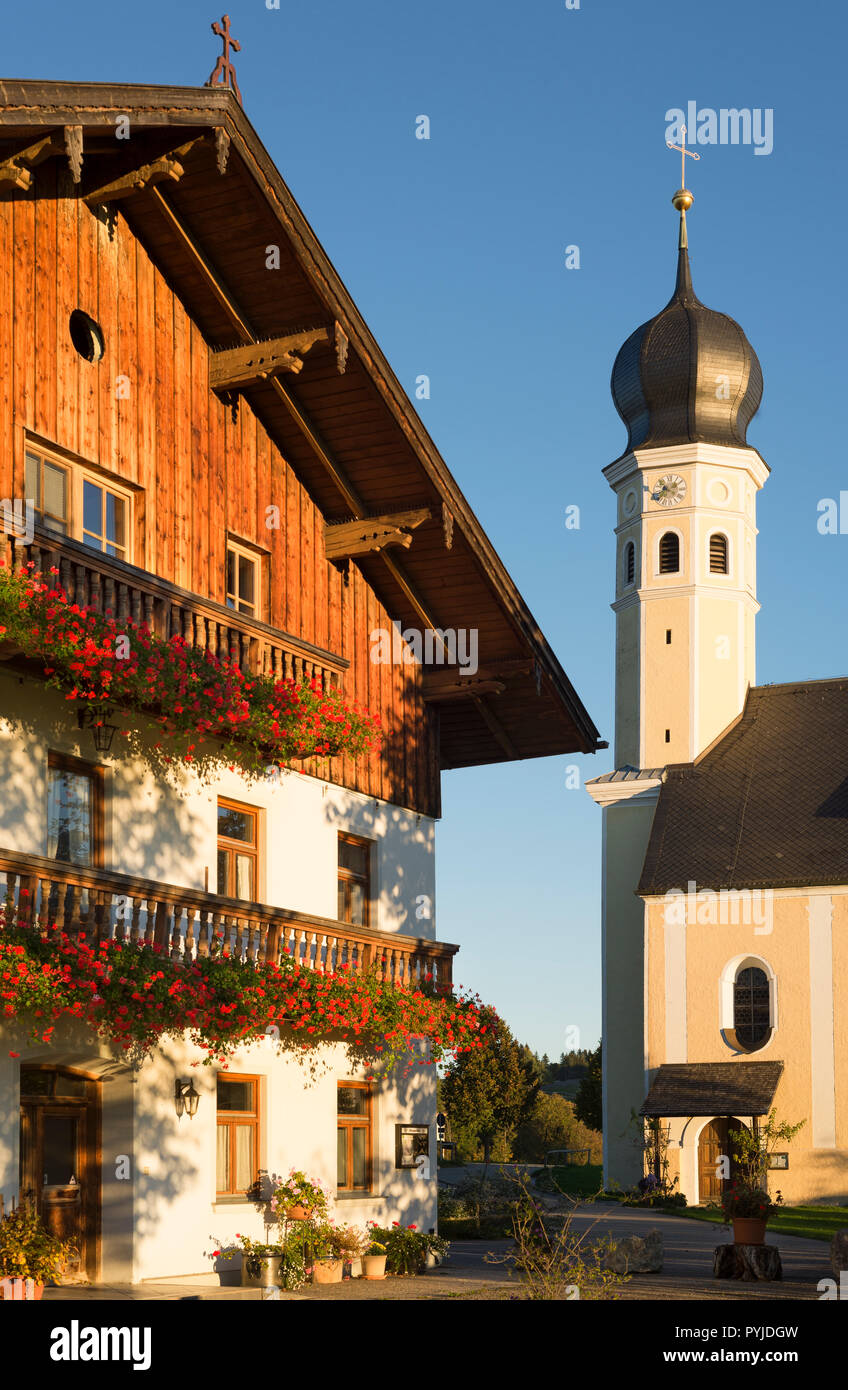 Church tower of the baroque pilgrimage church Wilparting and facade of a tavern with blooming geraniums on wooden balconies, Bavaria, Germany Stock Photo